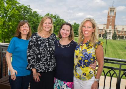 Four female alumni stand on the balcony and smile with the bell tower in the background. 