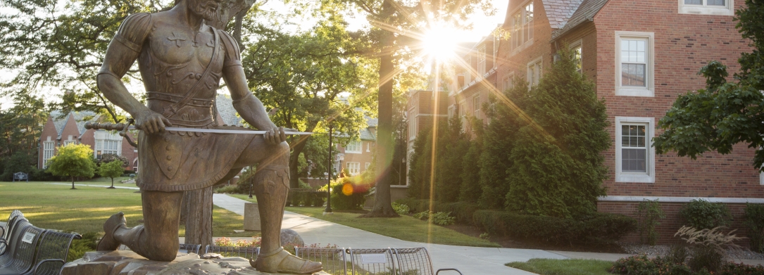 St. Ignatius statue on campus with sun shining through trees over building