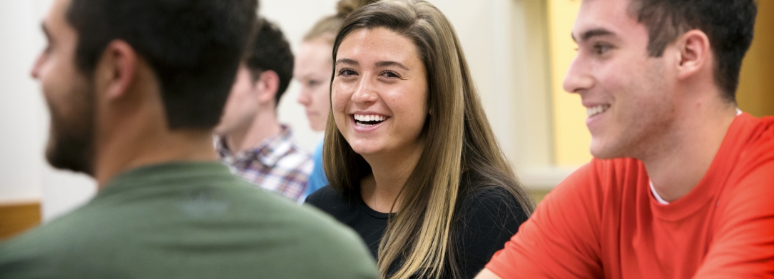 Three students sit in a classroom. One female student smiles at the camera.